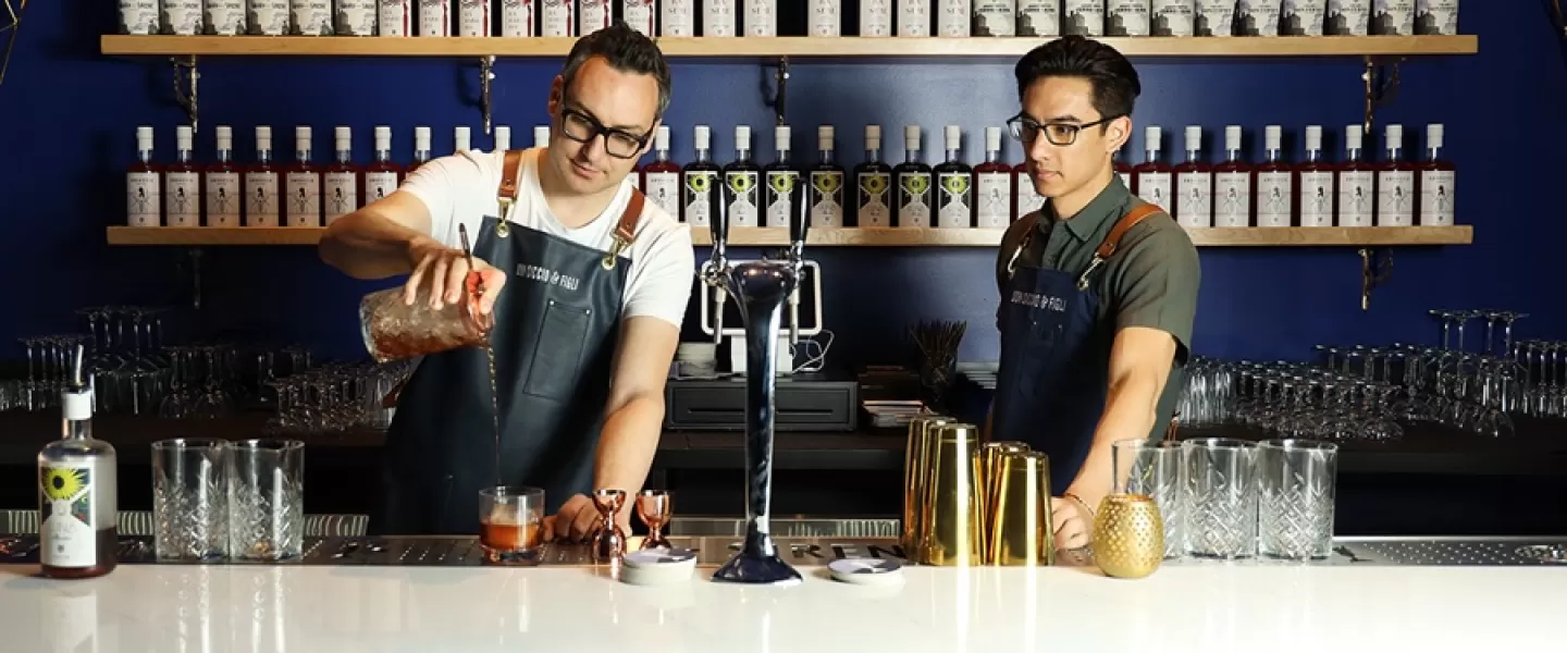 a male bartender with glasses and an apron pours a cocktail while another looks on, with shelves of liqueurs in the back 