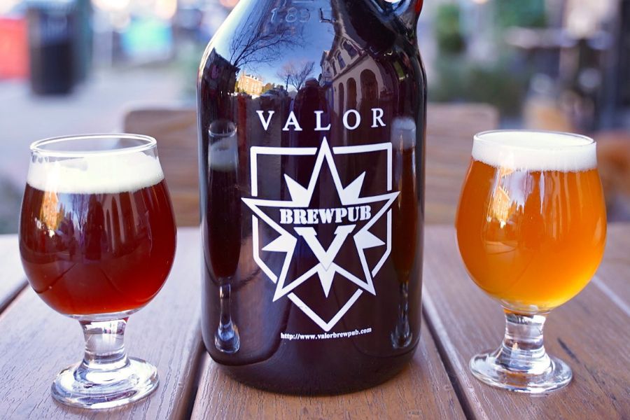 A growler and two glasses of beer on an outdoor wooden table, bearing the logo of Valor Brewpub. The beers are amber and pale gold in color