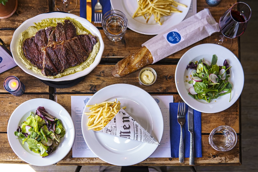 Overhead view of a table set with dishes including a large steak with sauce, fries in a paper cone, a baguette, and two salads, accompanied by drinks.