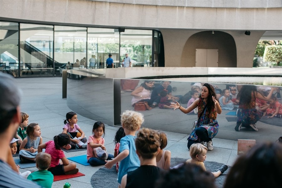A group of children seated around a storyteller in an open-air courtyard with a modern architectural background.
