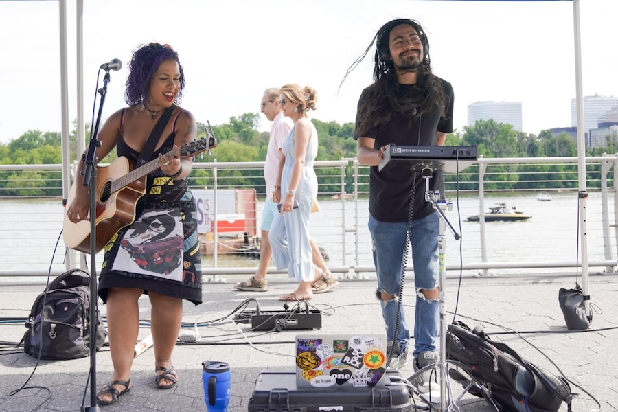 Two musicians perform outdoors by a waterfront. The female artist on the left plays an acoustic guitar while singing into a microphone. The male artist on the right plays a keyboard and smiles, with a background of pedestrians and a scenic river view.