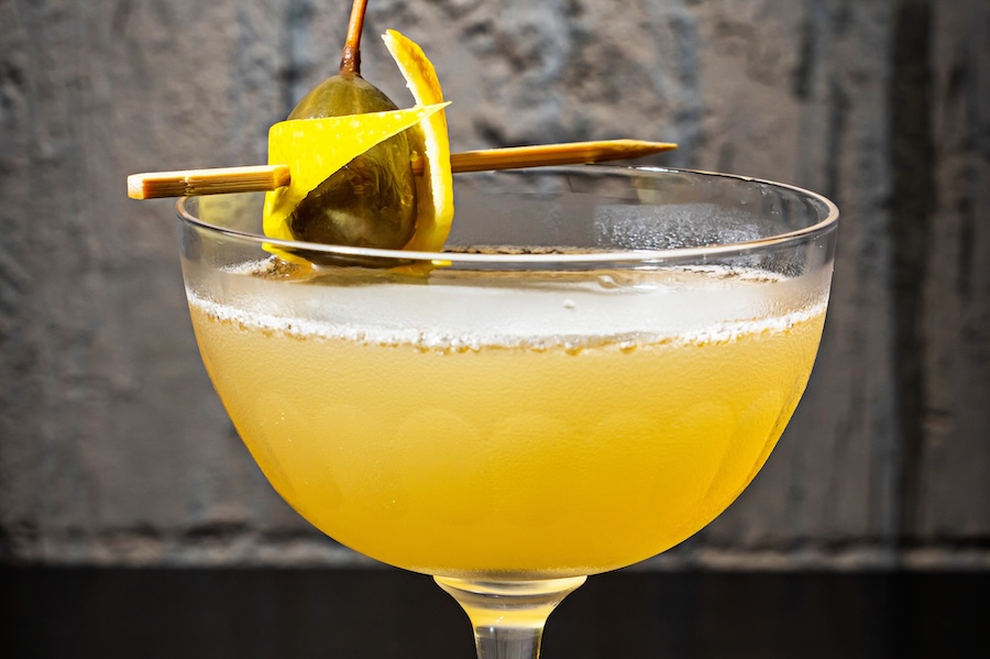 Golden yellow cocktail with an olive and lemon rind against a stone backdrop.