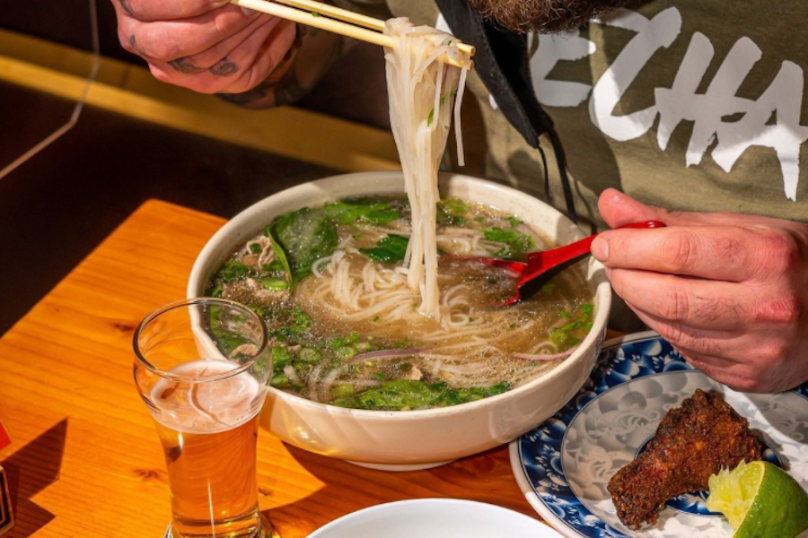 A person lifting noodles from a large bowl of pho with chopsticks, with a glass of beer and a piece of fried food on a plate nearby.