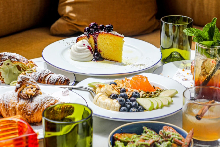 Close-up of a table set with various dishes, including a slice of cake topped with berries and whipped cream, pastries, a fruit and nut bowl, and assorted drinks in colorful glasses.