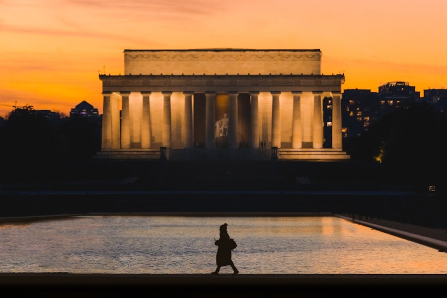 Die Silhouette einer Person, die am reflektierenden Pool entlanggeht, ist vor dem beleuchteten Lincoln Memorial bei Sonnenuntergang zu sehen, mit einem warmen orangefarbenen Himmel und Stadtgebäuden im Hintergrund.