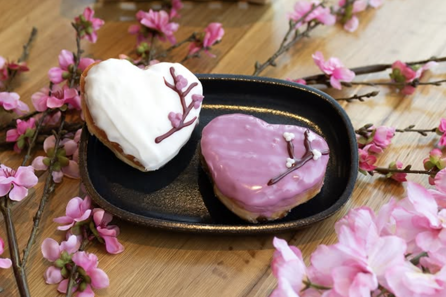 Dois donuts em formato de coração decorados com ramos de flores de cerejeira estão sobre uma mesa enfeitada com flores de cerejeira.