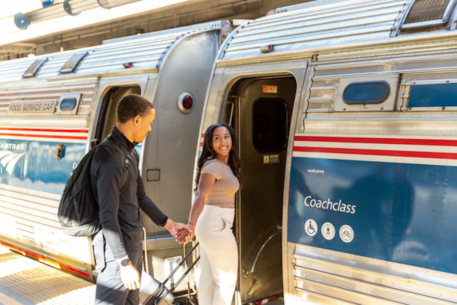 A couple smiles as they board an Amtrak train, holding hands and carrying luggage.