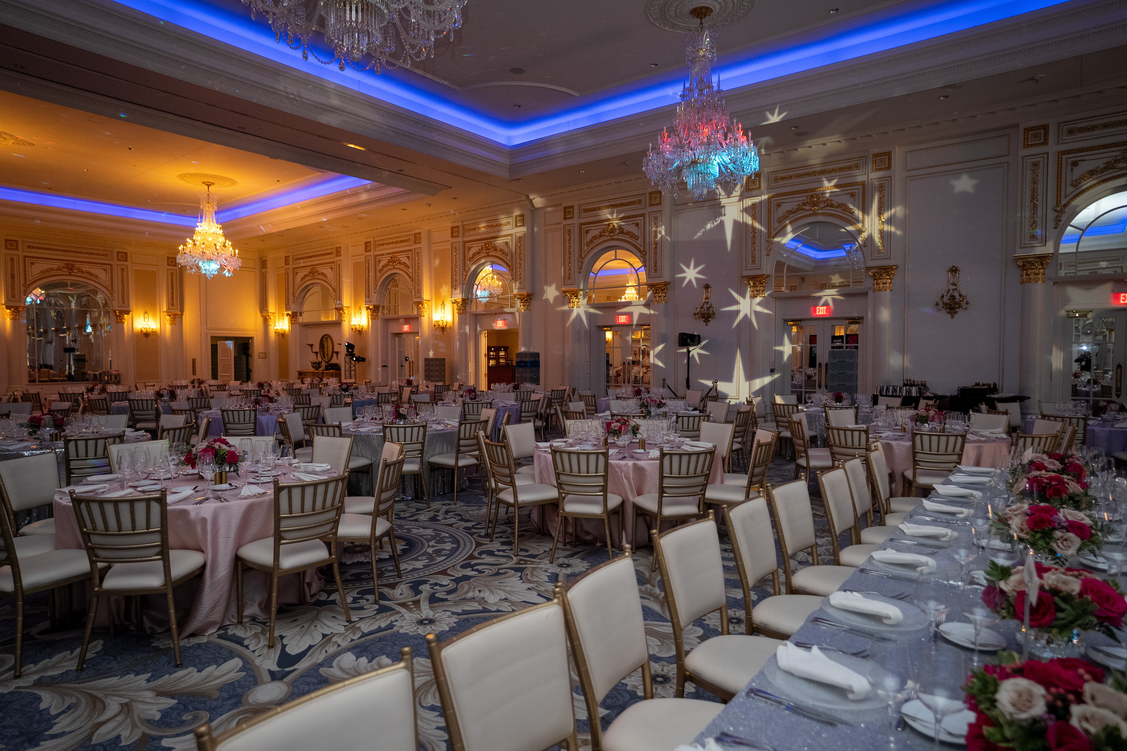 An elegant ballroom set for a formal dinner, featuring chandeliers, star-shaped light projections, round tables with pink linens, and floral centerpieces.