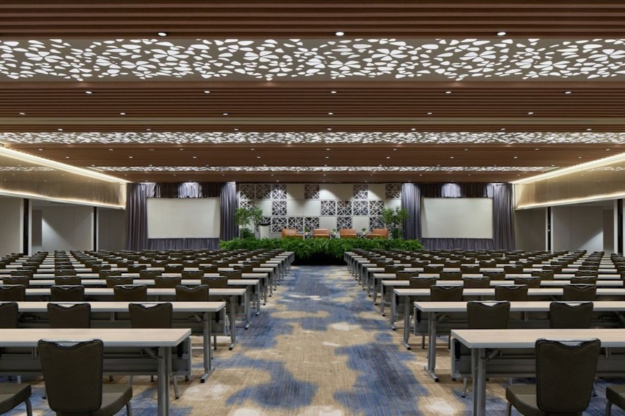 A large, modern ballroom set up for a conference with rows of tables and chairs facing a stage and projection screens.