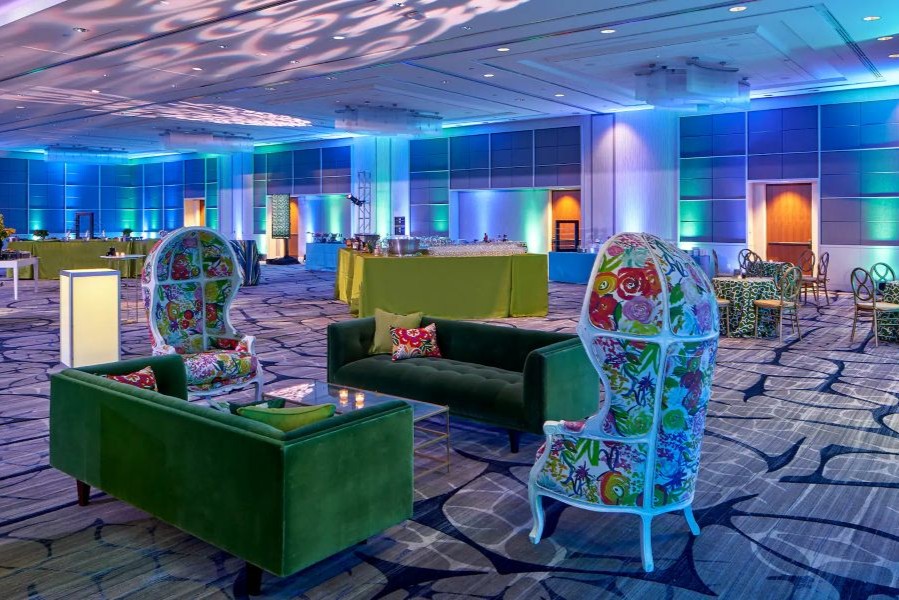 A colorful ballroom event setup featuring green velvet sofas, floral high-back chairs, cocktail tables, and vibrant blue and green accent lighting.