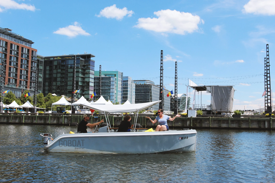 A small group enjoys a GoBoat rental on the water at The Wharf in Washington, DC, with modern buildings and tents along the pier.