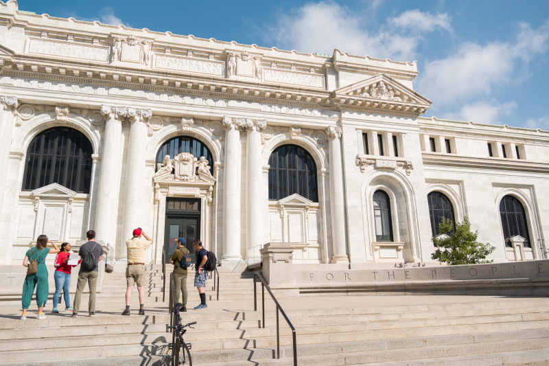 Visitors stand on the steps of the DC History Center, a grand marble building with tall columns and arched windows.