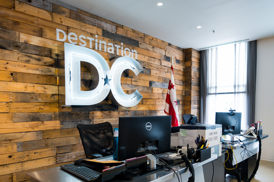 A front desk inside the Destination DC office with computers, a wooden accent wall and a large illuminated “DC” sign in Washington, DC.