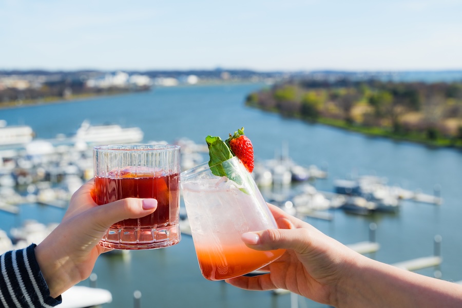 Two hands clink together colorful cocktails against the backdrop of a sunny day at the Wharf's waterfront.