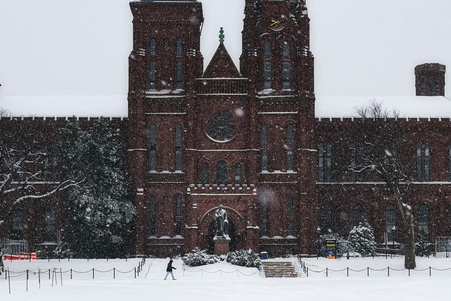 Snow falls heavily around the Smithsonian Castle as a pedestrian crosses the white-covered National Mall.