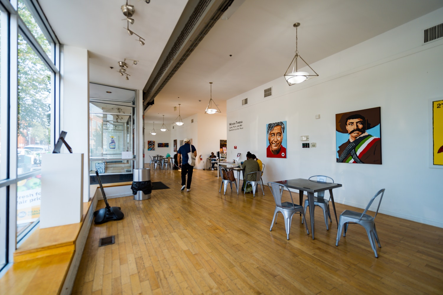 A bright, open gallery inside Anacostia Arts Center in Washington, DC, features local artwork on white walls, wooden floors, and café-style tables where visitors sit and work.