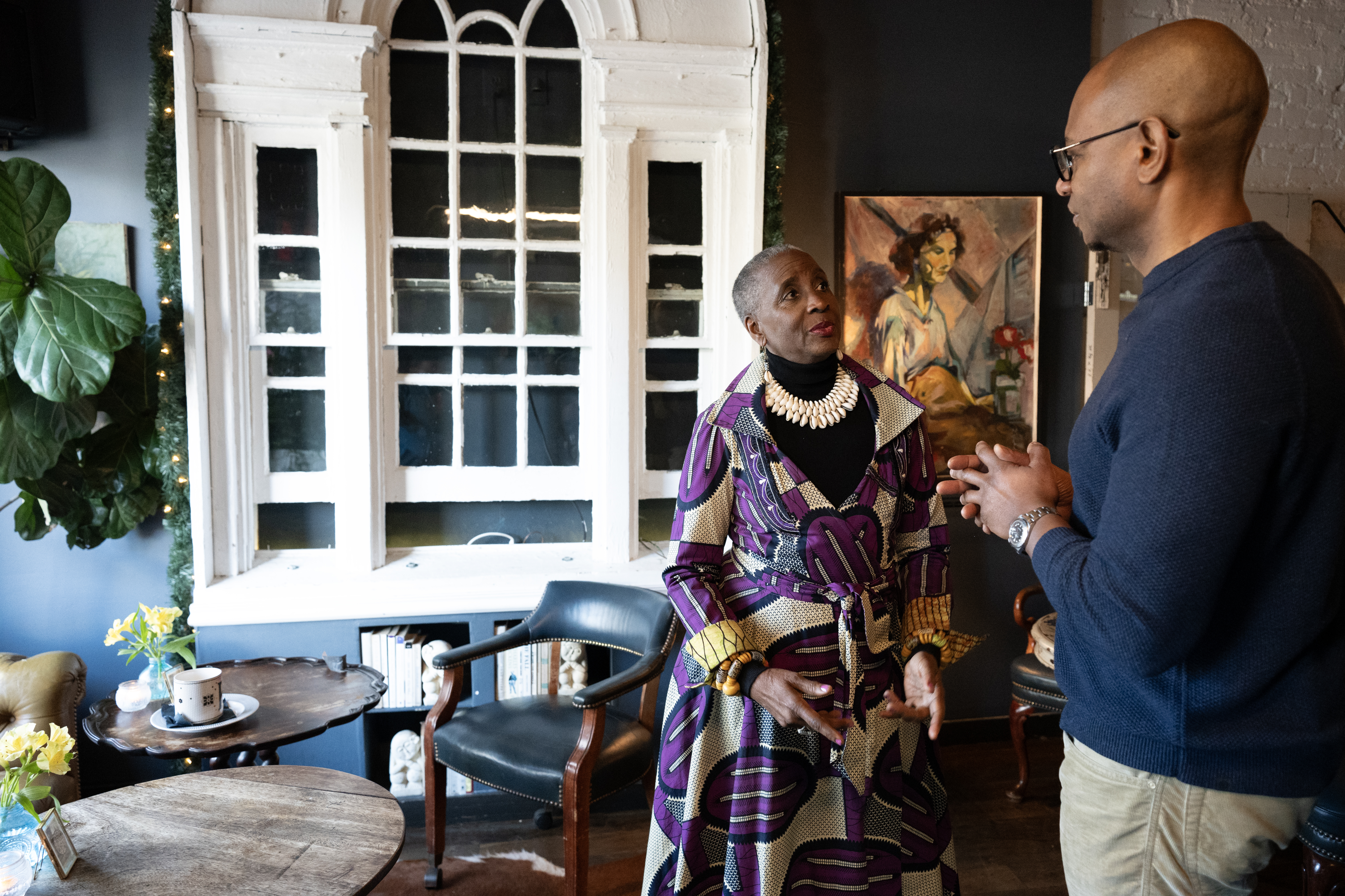 Two people have a conversation in an art-filled lounge with large windows and vintage furniture in Washington, DC.