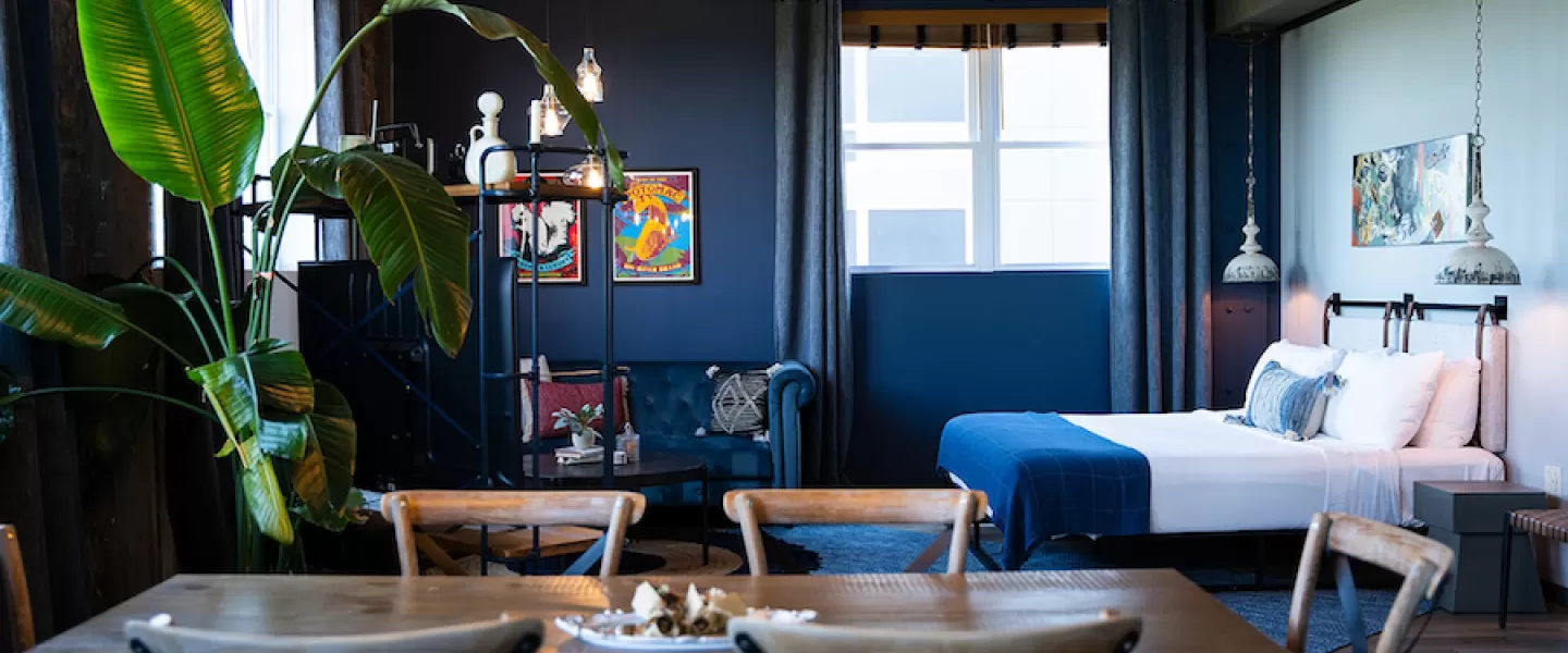 Cozy hotel suite with a dining table, large potted plant, blue sofa, and bed with blue bedding. Dark blue walls and framed artwork add color.