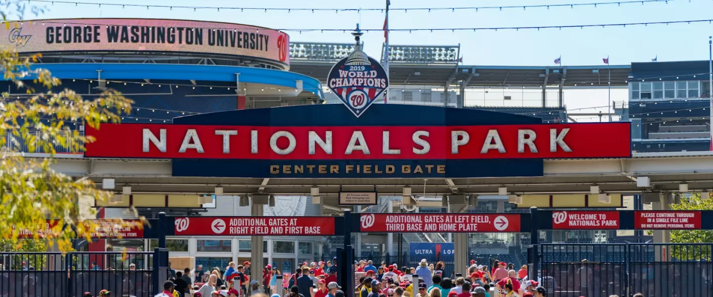 Crowds of fans dressed in red gather at the Center Field Gate of Nationals Park in Washington, DC, with a large "2019 World Champions" sign above the entrance.