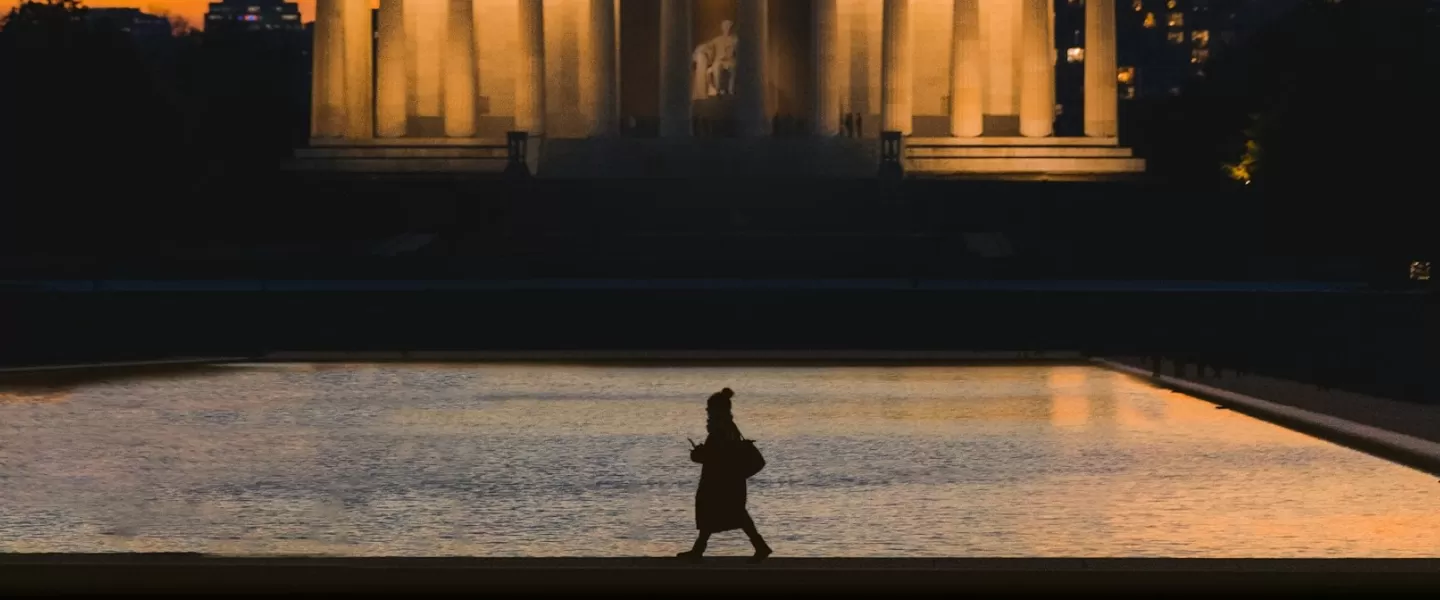 A silhouette of a person walking along the reflecting pool is set against the illuminated Lincoln Memorial at sunset, with a warm orange sky and city buildings in the background.