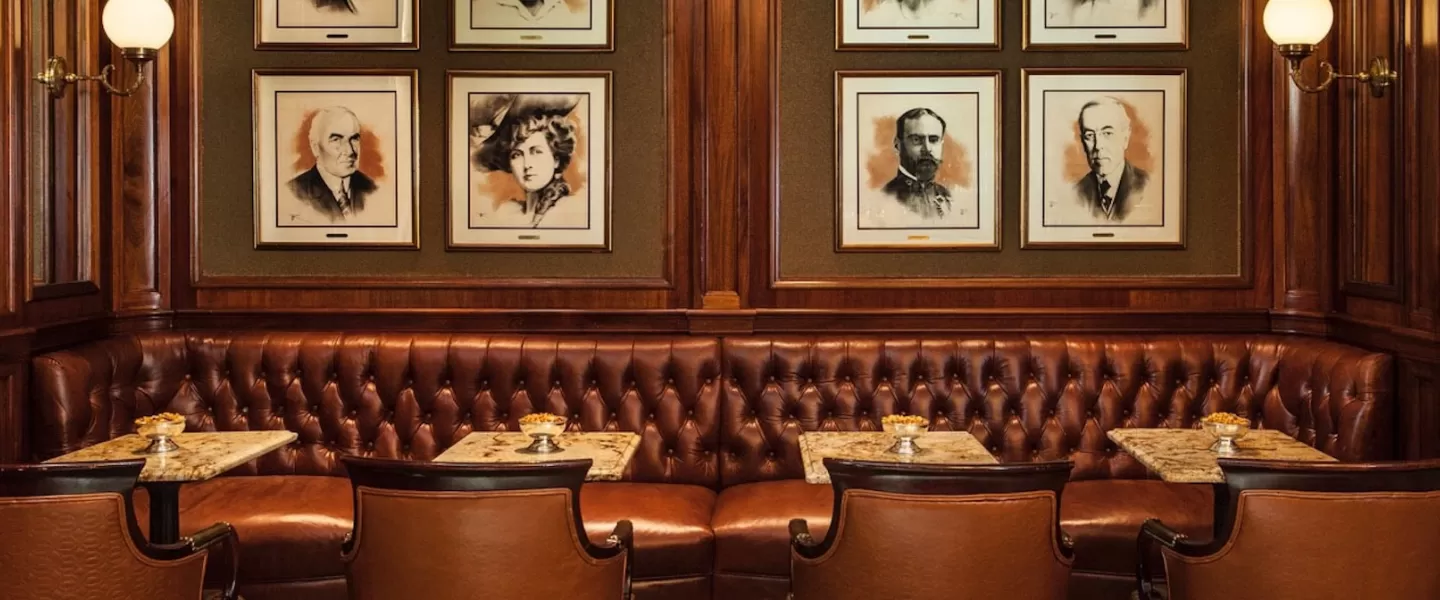 A cozy seating area featuring tufted leather banquettes, small marble tables, and portraits of notable historical figures adorning the walls in a warm wooden setting.