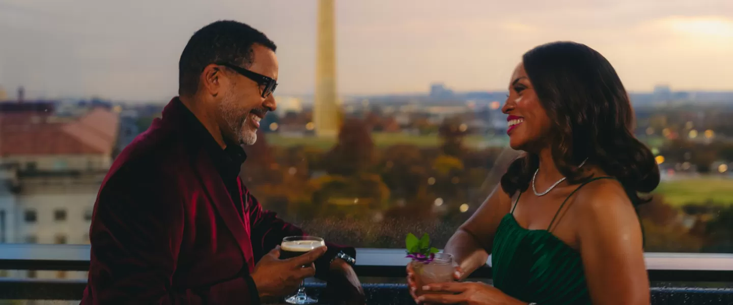 A well-dressed couple enjoys cocktails at The Vue rooftop restaurant with the Washington Monument in the background at sunset in Washington, DC.