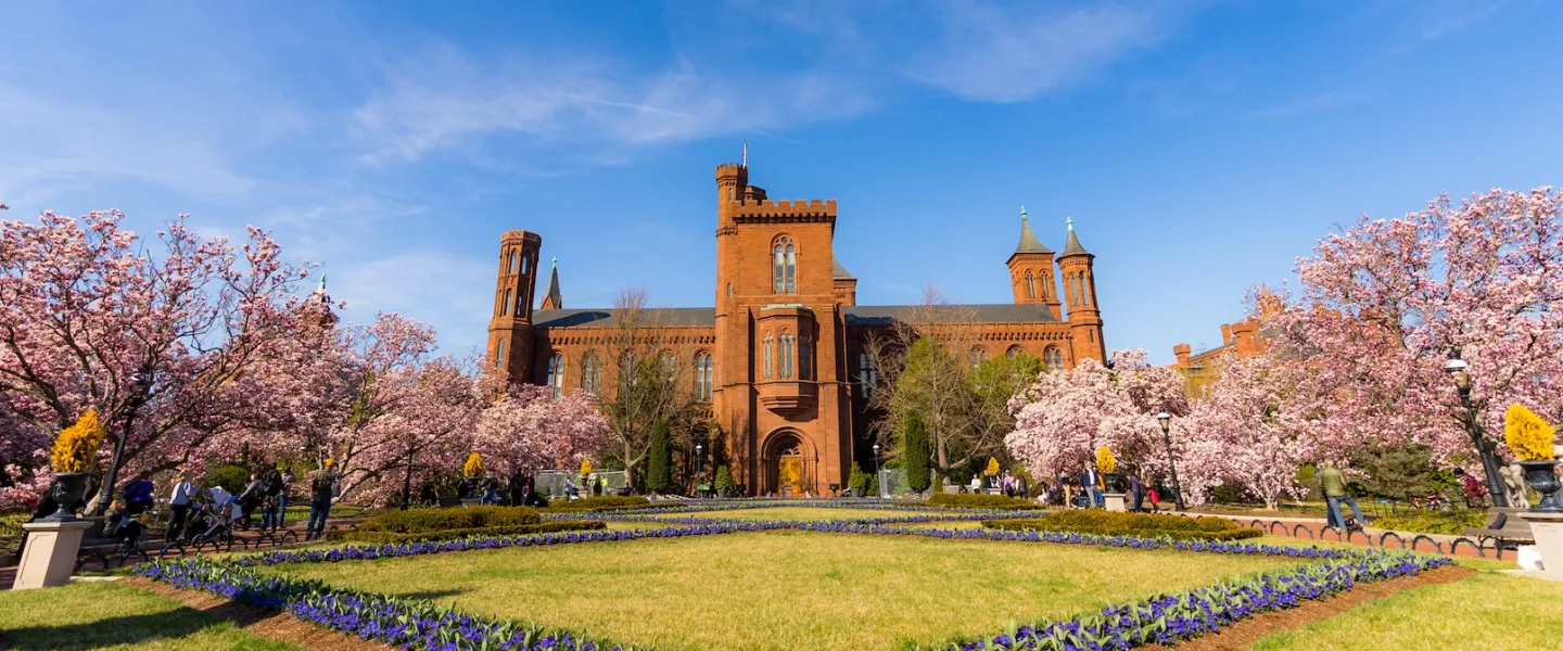 Visitors walk across the National Mall toward the red sandstone Smithsonian Castle on a sunny day.