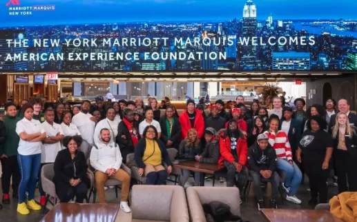 A group of high school students of color stand with General Manager Dan Nadeau and other hotel leaders in front of a large screen with the New York skyline and the word "New York Marriott Marquis Welcomes The American Experience Foundation

