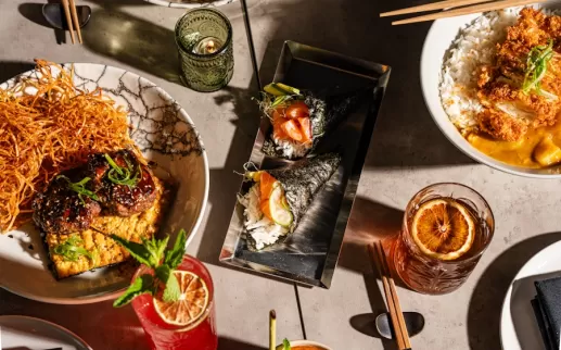 An overhead view of a diverse spread of Japanese dishes, featuring sushi rolls, rice bowls, and various appetizers, along with vibrant cocktails and meticulously plated dishes on a modern table setting.