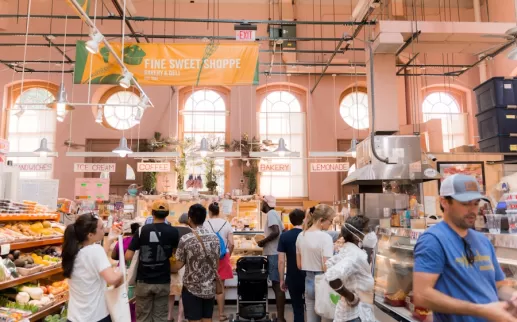 Shoppers line up at a bakery stall inside Eastern Market. The bright and airy space is filled with natural light, with signs for coffee, ice cream, and bakery items hanging above.
