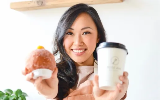 A smiling woman holds up a fluffy, filled donut in one hand and a coffee cup in the other.