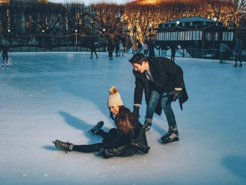 An ice skating rink with a woman who has fallen but is laughing, and a man laughing and helping her up.