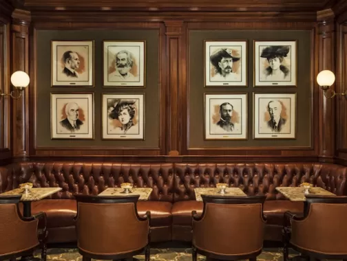 A cozy seating area featuring tufted leather banquettes, small marble tables, and portraits of notable historical figures adorning the walls in a warm wooden setting.