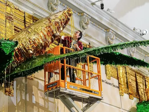 Artist installs a large gold and green piñata-style artwork from a lift inside a grand gallery.