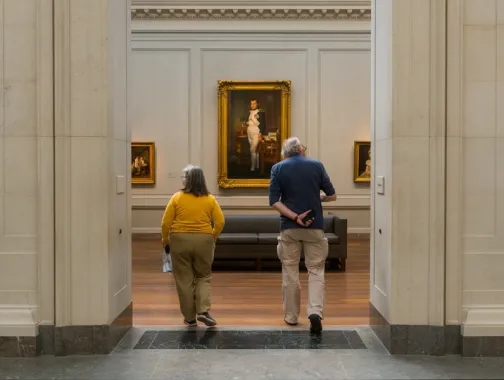 Two people walk into a museum gallery toward a large framed portrait hanging on the far wall.
