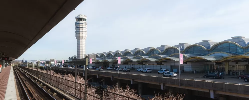 View of the exterior of Ronald Reagan Washington National Airport with the air traffic control tower and Metro tracks in the foreground on a clear day.