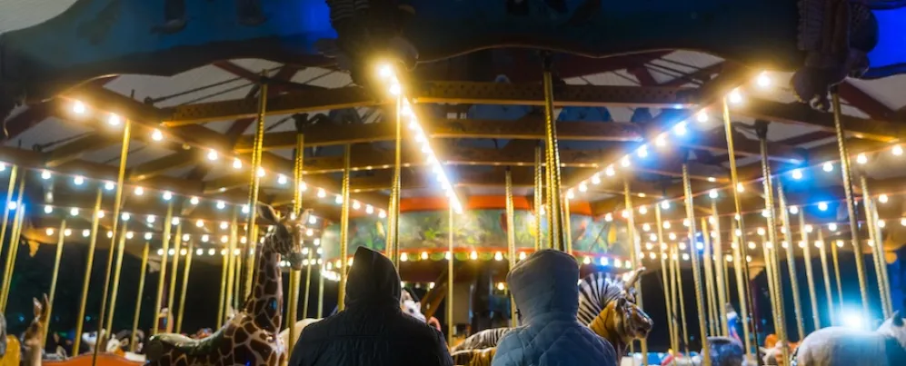 Two people in winter coats watch an illuminated carousel with animal figures during ZooLights at night.