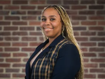 A person with long blonde braids wearing a dark blazer with a plaid pattern, smiling in front of a brick wall.